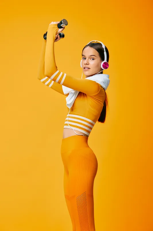 Fit young woman exercising with dumbbells in vibrant orange activewear, wireless headphones and towel, studio sport portrait on matching bold orange background.