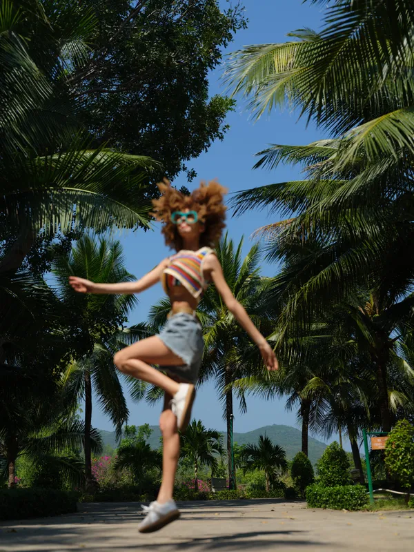 young woman jumping outdoors in tropical park, wearing colorful striped top and denim shorts, bright sunny day with clear blue sky and palm trees