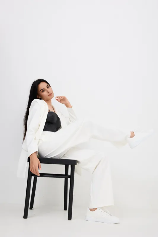 Sitting woman in white suit and black top poses confidently on black chair against plain white background, showcasing modern style and casual elegance.