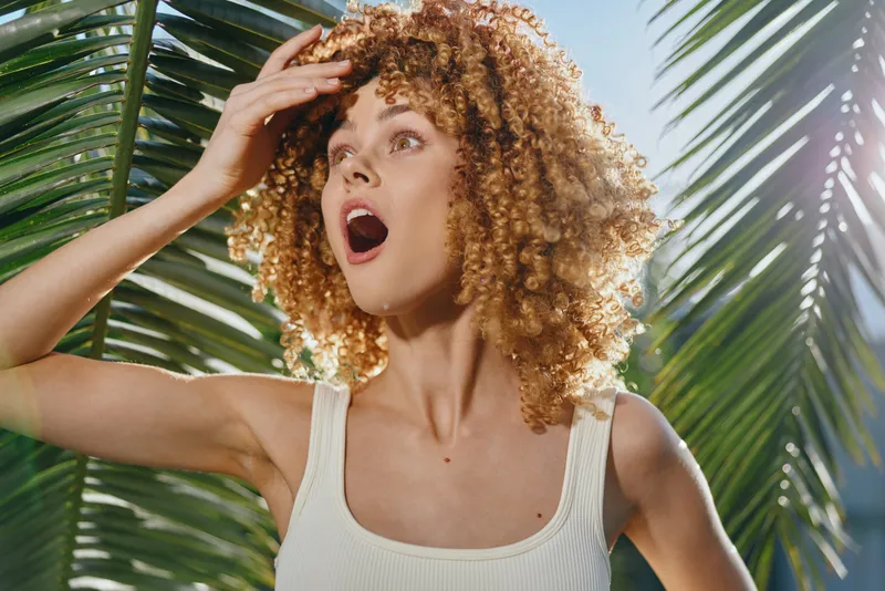 surprised woman with curly hair wearing white tank top stands outdoors among palm leaves under bright sunlight expressing amazement and wonder