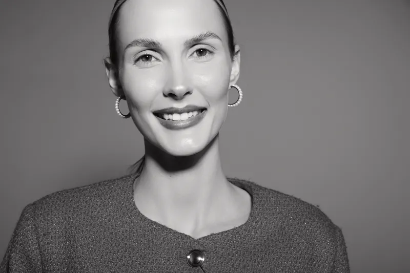 Smiling white woman with light skin and pulled back hair wears hoop earrings and a textured sweater. Studio portrait in black and white with soft lighting and clear facial expression.