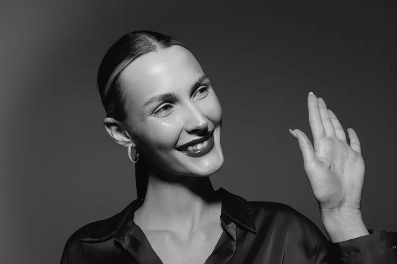 Smiling Caucasian woman with pale skin waves hand. Black and white portrait in studio with soft lighting. Concept of greeting, elegance, and female beauty.
