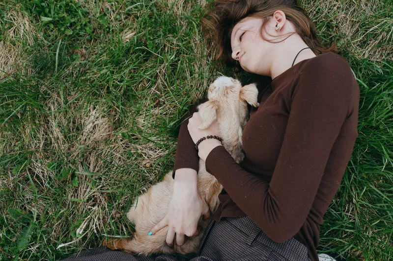 Candid woman outdoors, cuddling a small dog in a warm embrace, brown tones, lush grass background, peaceful moment, casual style, natural light, authentic lifestyle scene