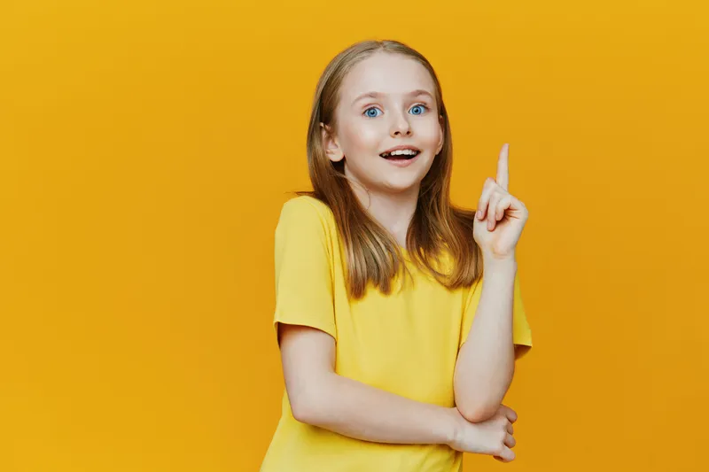 Cheerful young girl with light hair in a yellow shirt on a bright yellow background, expressing excitement and creativity through her joyful gesture