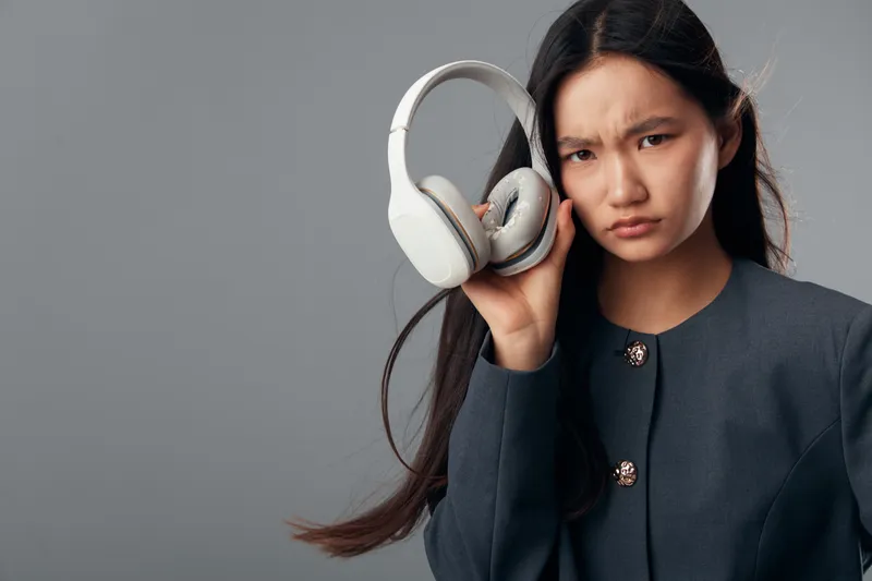 Woman holding headphones close to her ear with a confused expression, wearing dark jacket on grey background. Studio portrait of female listening with doubt and uncertainty.