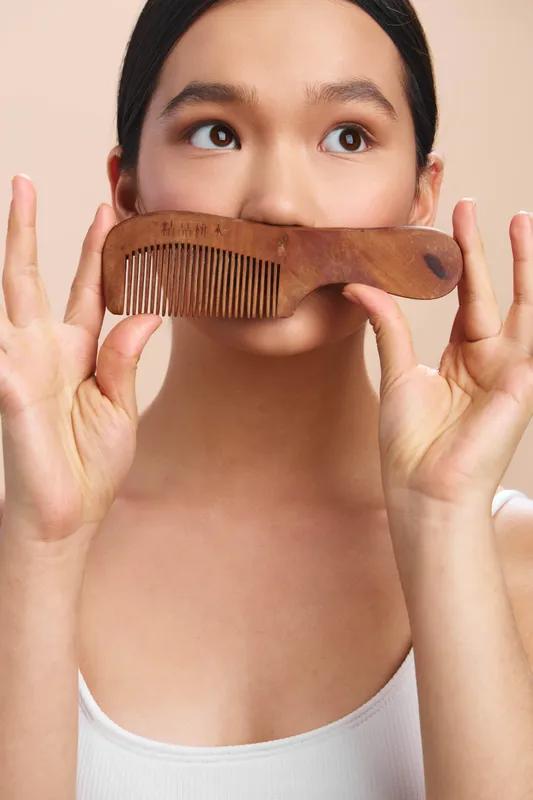 asian woman holds a wooden comb near her face in a closeup portrait, playful expression, soft lighting and simple background creating a casual fashion mood.