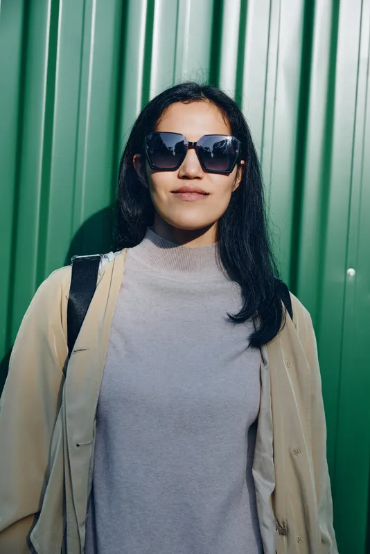 Woman in dark sunglasses leans against a green wall, dressed in a gray turtleneck and beige coat, showcasing chic street style and calm confidence in urban setting.