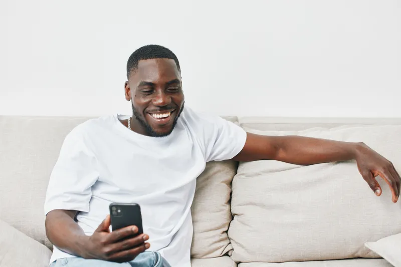 Happy young African American man enjoying his smartphone on a grey couch, smiling while sitting casually in a bright living room