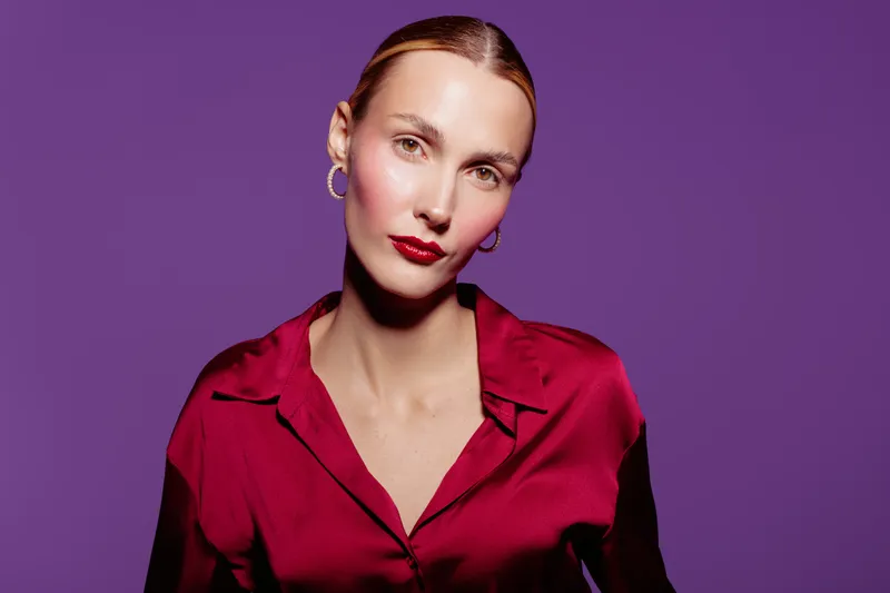 Portrait of confident woman with slicked hair wearing red blouse and hoop earrings on purple background. Studio lighting highlights her makeup and serious expression.
