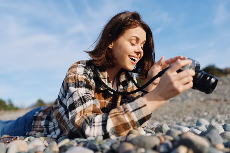 Young photographer lying on a rocky beach, smiling, capturing vibrant moments with a compact camera under a bright, clear sky and rolling pebbles