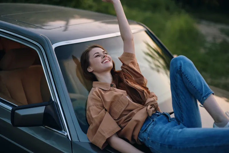Young woman enjoying a joyful moment, reclining on a vintage car during a sunny day, wearing a casual outfit with a vibrant smile, conveying a sense of freedom and happiness.