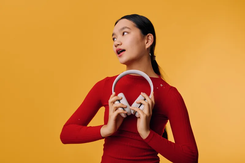 Young woman in red sweater holding white headphones looking aside with thoughtful expression on yellow background. Portrait of girl with dark hair in casual style for music and technology.