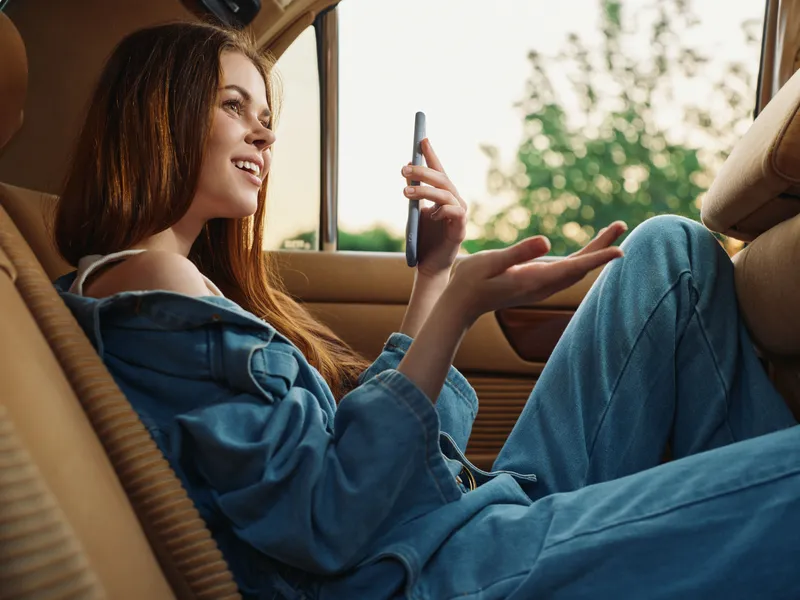 Young woman in denim outfit smiling while talking on phone in a car, capturing a moment of joy and connection in a serene setting.