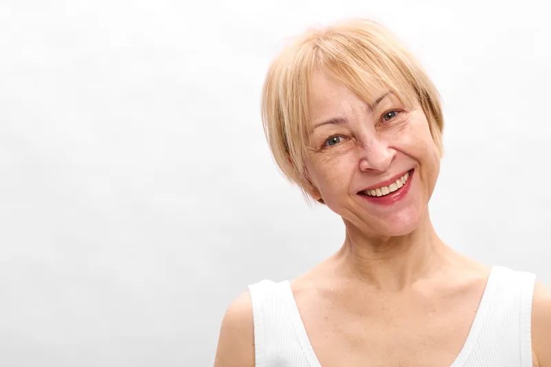 Woman portrait, smiling blonde mature woman with short hair, studio background, gentle light, white top, relaxed posture, soft neutral colors, copy space
