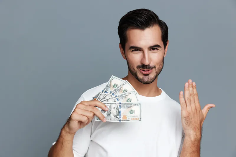 Confident man holding banknotes in hand smile at camera wear casual white t-shirt posing on gray background studio portrait. Money success concept.