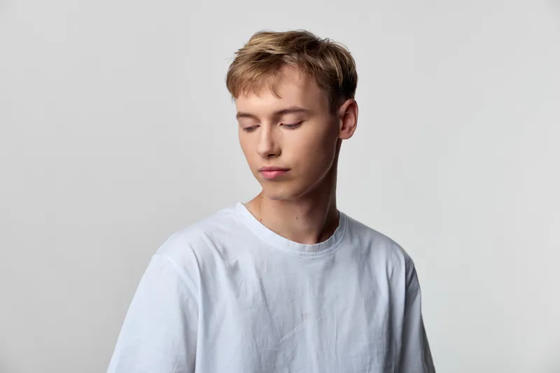 Young man in a white shirt stands in a studio with a calm, contemplative mood, soft lighting and a minimal background for a clean fashion portrait