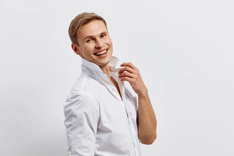Smiling man with light hair in white shirt looking at camera, cheerful expression, studio portrait on plain gray background, lifestyle concept, positive mood, casual attire, happiness, confidence, you