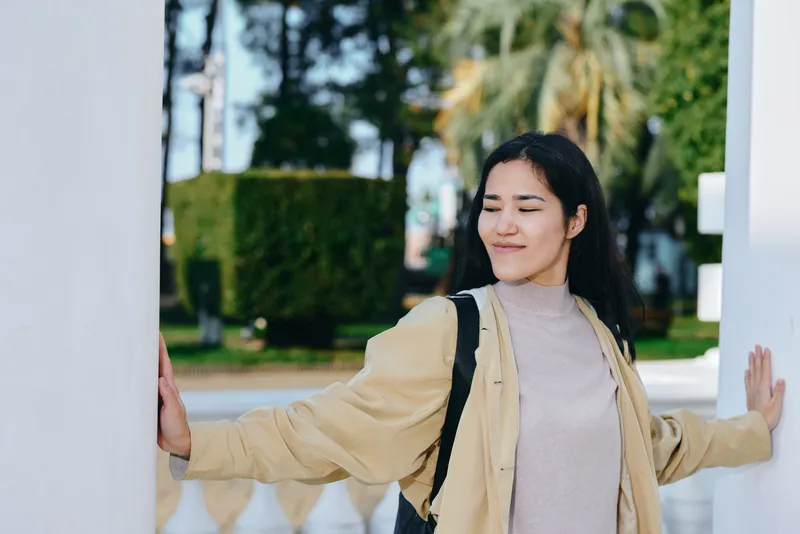Woman in casual jacket smiles while leaning on white columns outdoors, backpacked fashion, sunny day, featuring relaxed style and fresh campus vibe, and modern energy