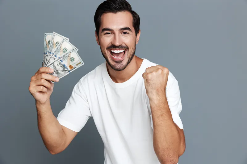 Happy smiling man holding money in hand, celebrating success and achievement in modern casual clothing against a neutral gray background. Lifestyle concept.