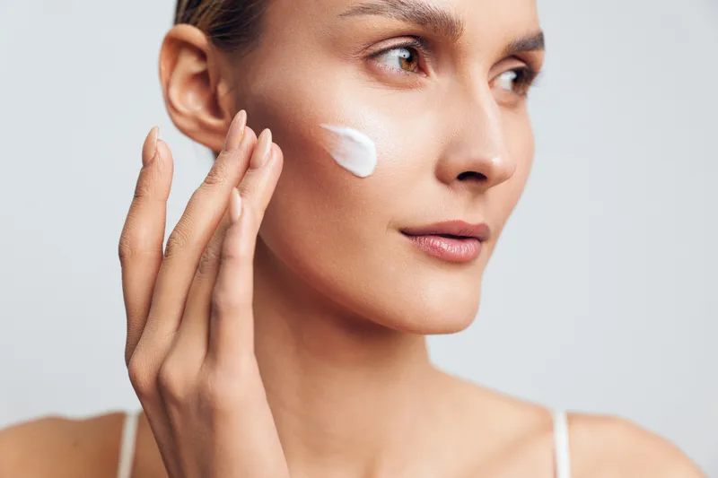 A young woman applies white cream to her cheek with fingers in a close-up portrait against a plain background. Concept for skincare, beauty, and facial care routine.