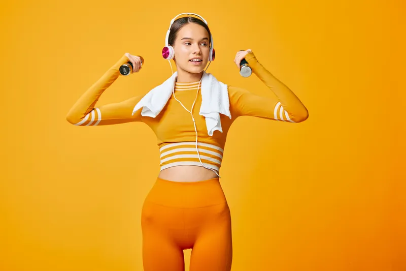 Young woman in orange athletic wear with headphones, lifting dumbbells while a towel rests on her shoulders in a vibrant studio. Energetic fitness routine, dynamic pose, and focused workout energy.