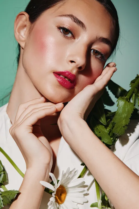 Close-up portrait of a young woman with glossy red lips and dewy skin posing creatively with hands near face and white flowers on mint background beauty fashion concept.