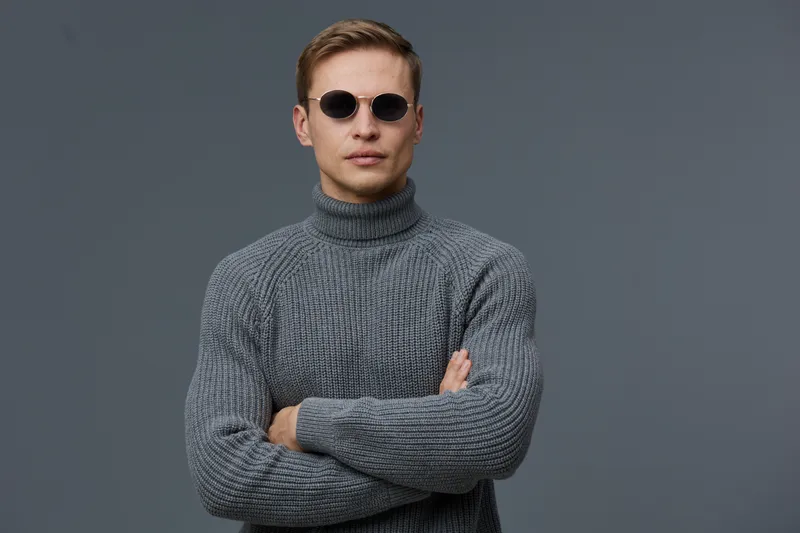 Man with stylish sunglasses wearing a gray turtleneck sweater stands confidently against a plain gray background, exuding coolness and modern fashion. Portrait male fashion neutral studio shot.