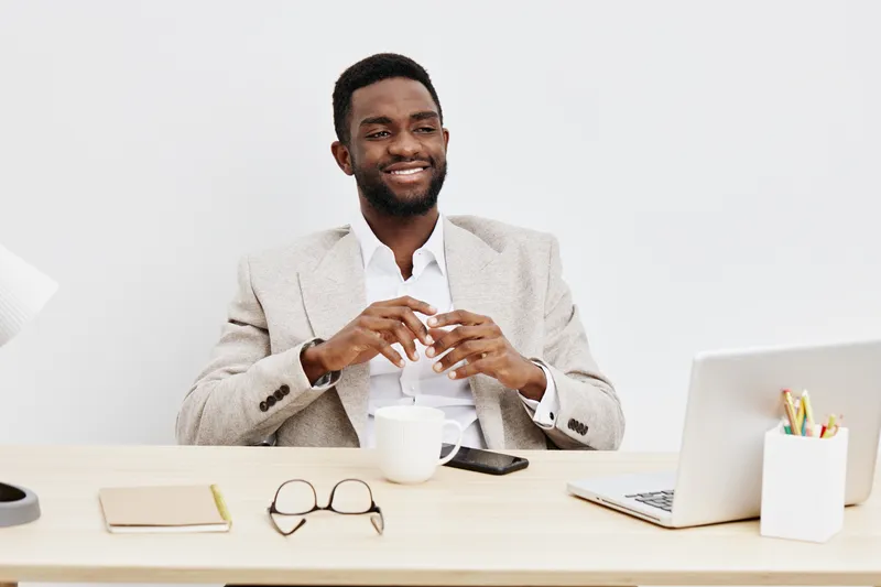 Smiling young African American man in light beige blazer sitting at modern office desk with laptop, smartphone, notebook, coffee mug, and glasses, happily looking at the camera, positive business life