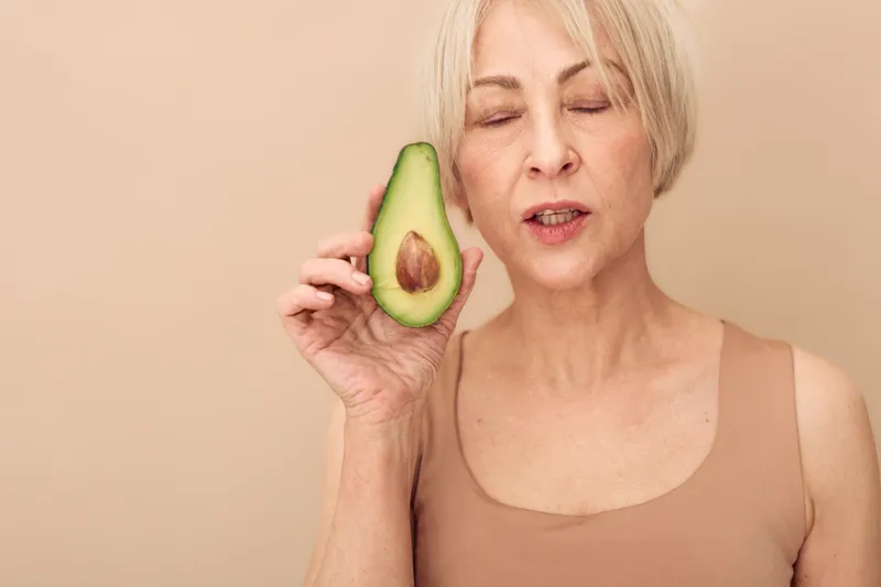 Mature woman with closed eyes holds fresh avocado half near face on beige background. Concept for healthy eating, natural skincare, wellness, and nutrition for middle aged adults.