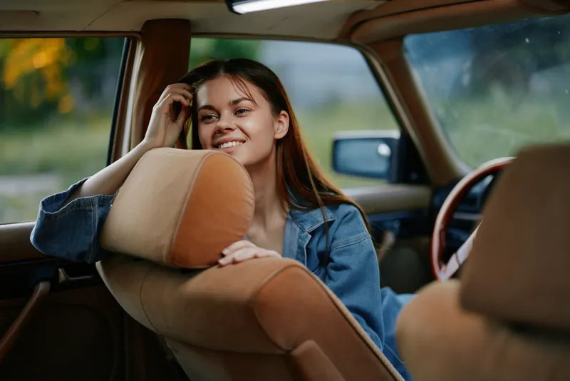 Young woman smiling in a vintage car, dressed casually in denim, exuding joy and relaxation against a warm and natural background, perfect for lifestyle photography.