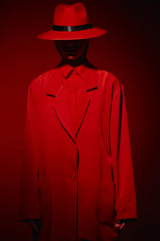 Fashion model dressed in red oversized blazer and wide-brimmed hat posed against intense red background. Creative beauty concept highlighting bold red clothing and mysterious style.