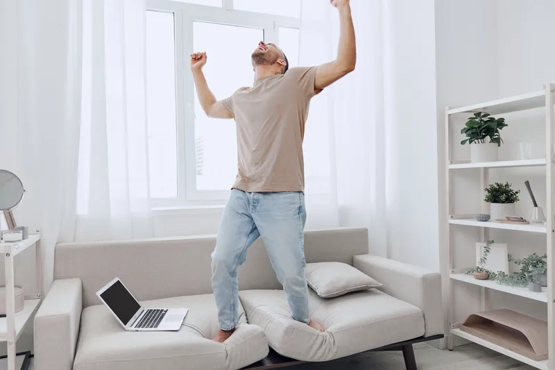 Energetic man celebrating success in a bright, modern living room, showcasing a joyful moment filled with light and positivity while working from home.