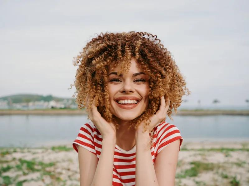 Cheerful young woman with voluminous curly hair in a red and white striped shirt smiles at the camera on a sunny beach with calm water in the background.