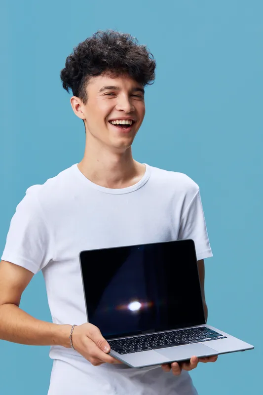 smiling man holding laptop with blank screen wearing casual white t-shirt against blue background, cheerful young adult with curly hair showing device