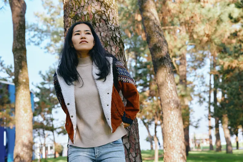 Woman outdoors in a park among trees, wearing a sweater and jacket, casual fashion and confident stance under sunlight, showcasing relaxed lifestyle in nature and scenery