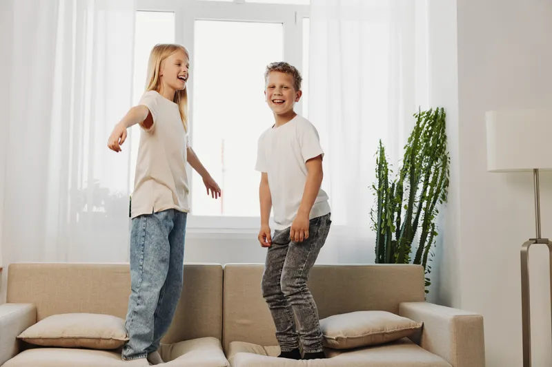 Two happy children jump on a beige sofa in a bright living room with a large window and green plant. Casual clothes and joyful mood emphasize carefree childhood fun.