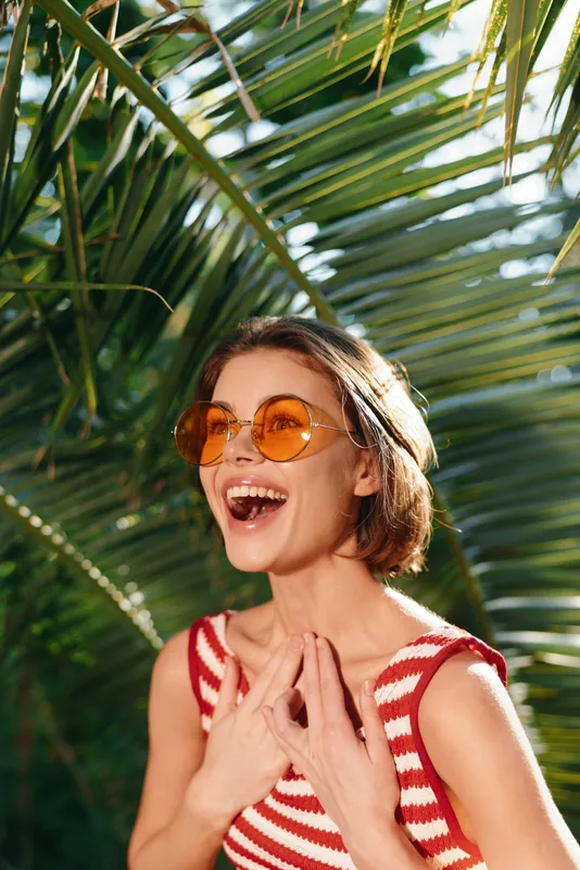 A joyful woman wearing round orange sunglasses and a striped red and white top stands outdoors among tropical plants, smiling widely and enjoying a sunny moment.