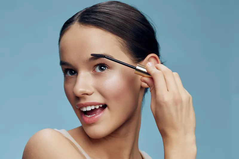 Young woman applying eyebrow gel, expressing joy, with a soft blue background, showcasing beauty, self care, and modern makeup trends