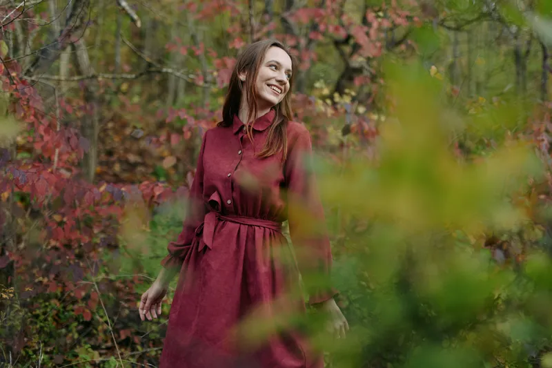 A smiling young woman in a simple burgundy dress stands among autumn foliage, conveying an authentic and genuine connection to nature, with a relaxed, candid portrait mood and warm tones.