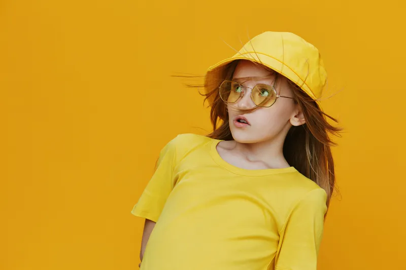 Young girl wearing a yellow outfit with sunglasses and a hat, posing against a vibrant yellow background, exuding a playful and cheerful vibe