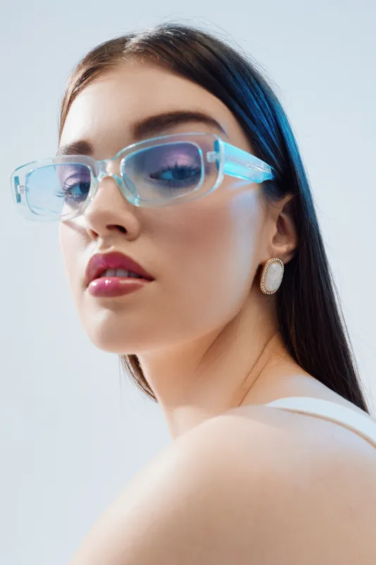 Young woman with long dark hair wearing transparent blue glasses and white earrings poses against a light background. Concept of beauty fashion and modern style.