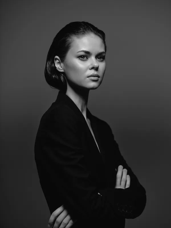 Portrait of a serious woman in black and white with crossed arms, displaying confidence and determination in a minimalistic studio setting.