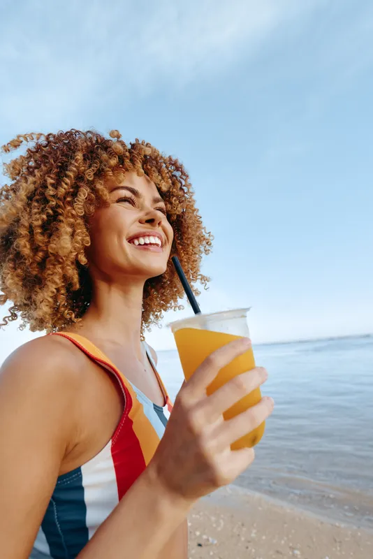 Curly hair woman smiling on beach, holding drink cup with straw, ocean waves behind, sunlight glinting, relaxed shoulders, warm summer day, carefree mood, refreshing vacation breeze