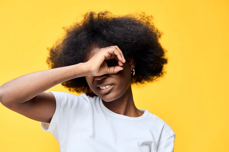 Young Black woman with natural curls smiling while playfully obscuring her face against a bright yellow background, conveying joy and confidence