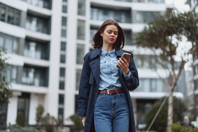 Woman in denim jeans and a light blue shirt checks her smartphone while walking outdoors. Urban architecture and street fashion create a casual, confident daily life mood.