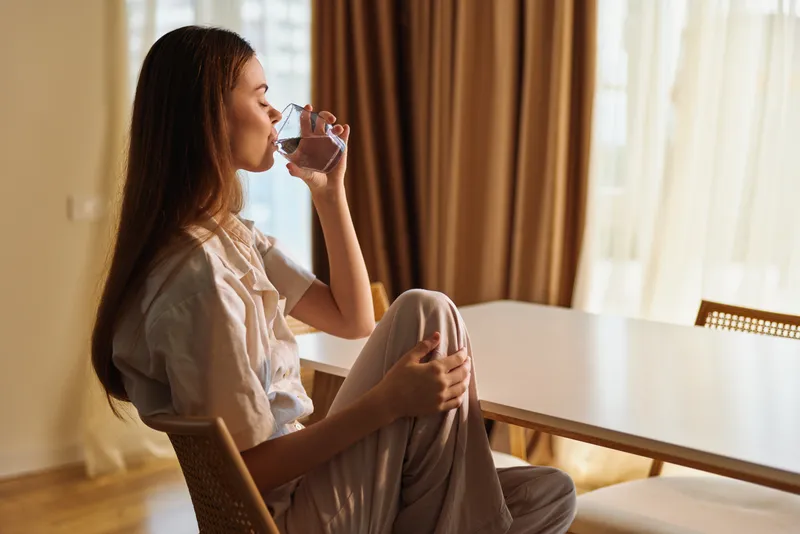 Woman sitting at a home table, drinking water from a glass in a relaxed morning scene with soft sunlight and curtains, casual portrait showing hydration, calm lifestyle moment.