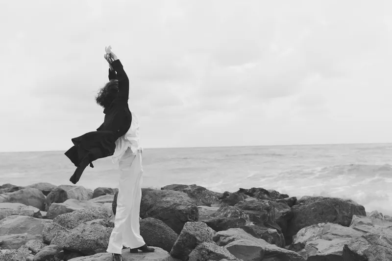Black and white scene of a couple on a rocky shoreline, one lifting the other in a joyful celebration by the sea, windblown clothes and a calm horizon.