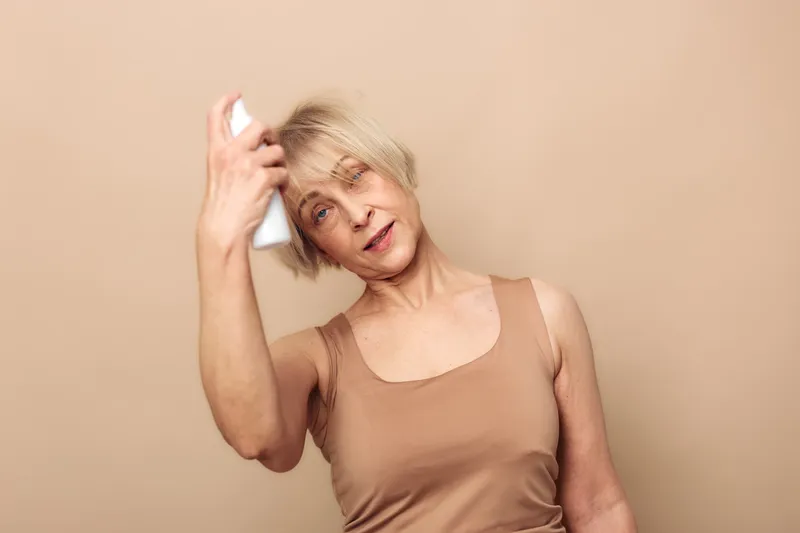 Mature woman sprays facial mist on her face with closed eyes in a beige tank top. Concept of skincare routine, hydration, self-care, and beauty for aging skin in a neutral studio setting.