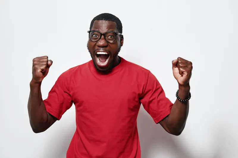 African American man with dark skin wearing glasses and red t shirt celebrating with fists up and mouth open on white background. Excited, happy, success and joy expression.