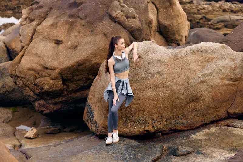 Young female athlete in stylish activewear sitting on a rock by the beach, embodying a sense of tranquility and fitness in nature, capturing the essence of a healthy lifestyle